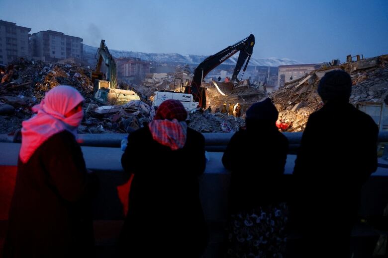 Black silhouettes of people seen from behind while they watch a digger in a lit-up pile of rubble.