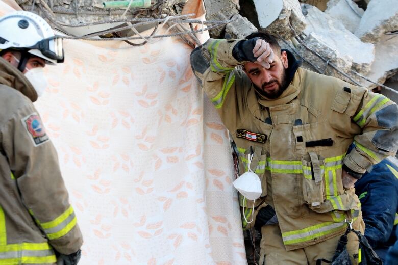 A man in a uniform, similar to a firefighter's, stands looking weary next to rubble.