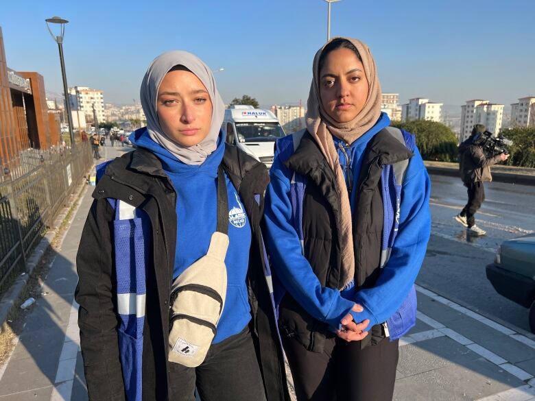 Two women wearing beige headscarves, blue sweatshirts, and black pants and jackets post for a picture on the sidewalk next to a street. One woman wearing a belt bag around her, while the other clasps her hands together. In the background, a blue sky and white buildings are shown.