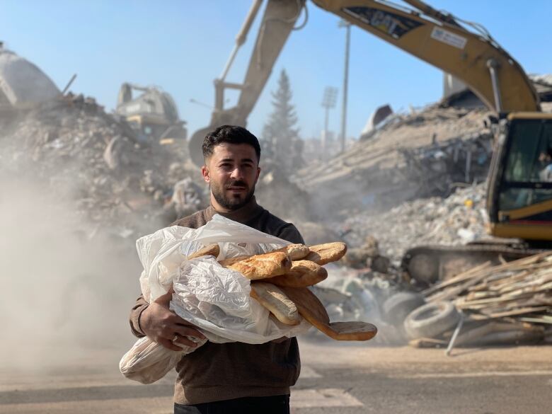 A man with dark hair, wearing a brown sweater, carries a plastic bag full of bread outside. Behind him, an excavator works to move rubble, casting up some dust.