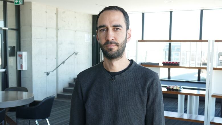 A man is standing in a brightly lit penthouse common room with large windows, tables, chairs and a pool table in the back ground.