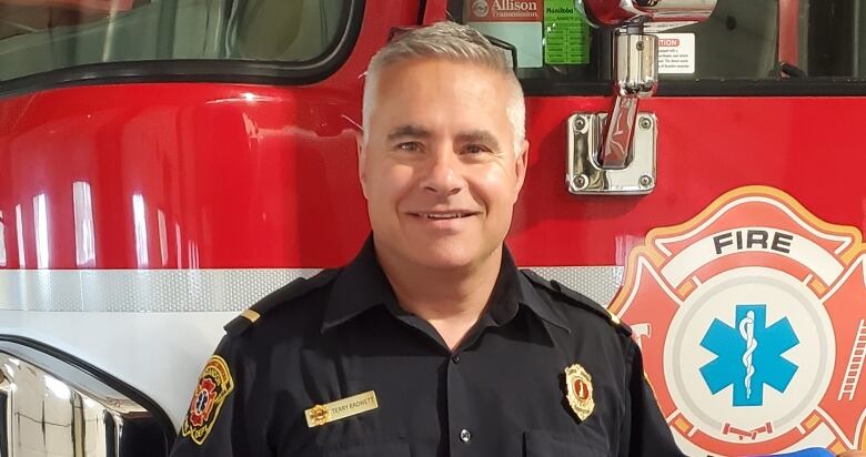 A man dressed in a firefighter uniform holds a lacrosse jersey in front of a fire truck.