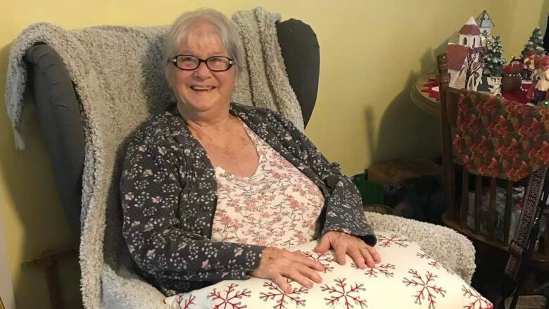 An elderly woman sits in a chair and smiles at the camera.