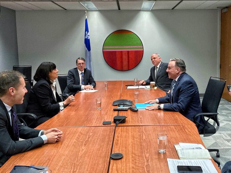 Five people sit around a long boardroom table, a single Quebec flag sits in the background