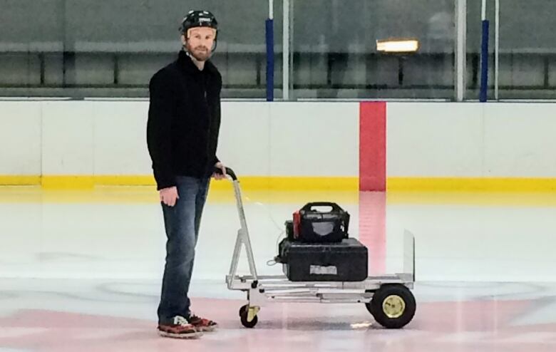 A man wearing a helmet pushes a metal cart carrying air monitoring equipment across the ice of an arena.