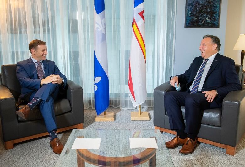 Two men sit on leather chairs spaced 2m apart. Behind them are the Quebec and Newfoundland and Labrador flags