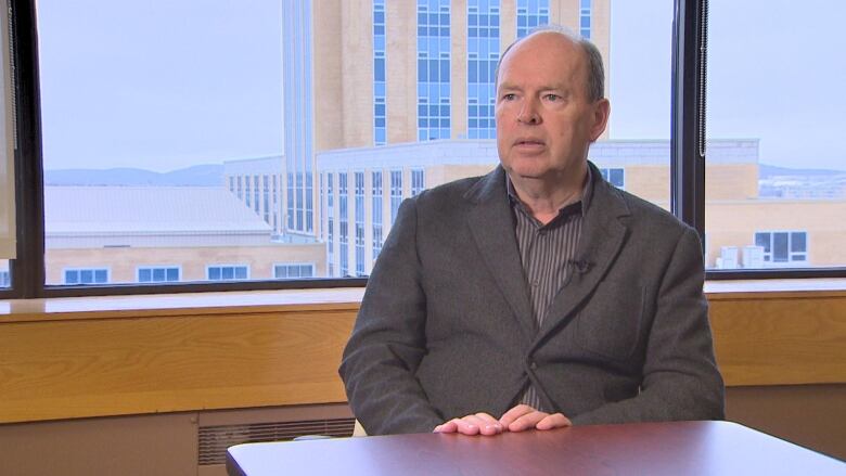 A man wearing a striped shirt and grey blazer sits at a table with the Confederation Building visible in the window behind him.