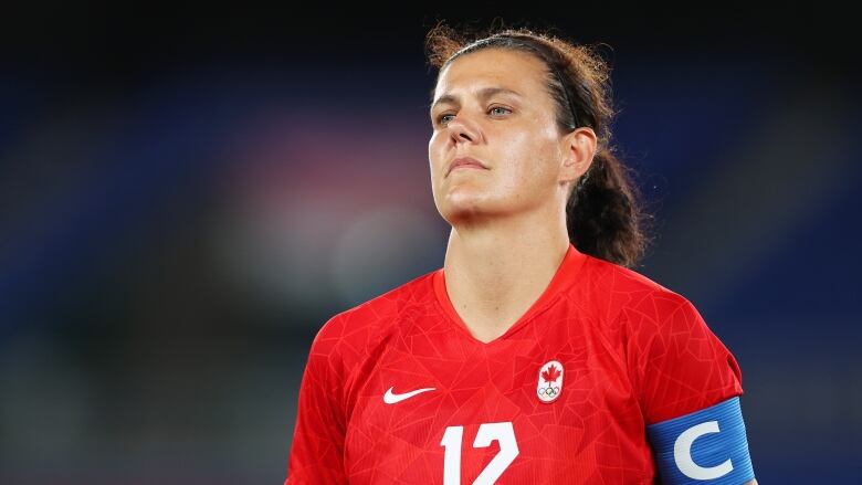 A soccer player in a red jersey stands with her hands behind her.