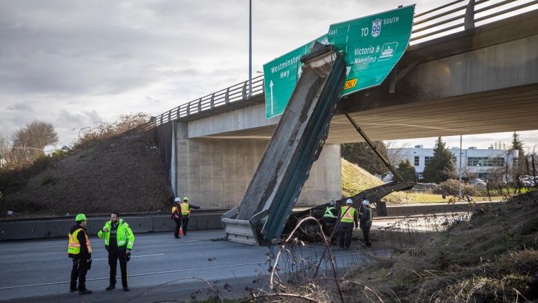 A commercial truck trailer is tilted up against an overpass above a highway. First responders are gathered on the ground.