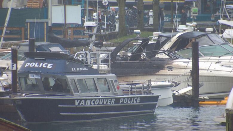 A blue boat marked 'Vancouver Police' sits moored at a dock next to a white boat which has clearly been burned and charred.