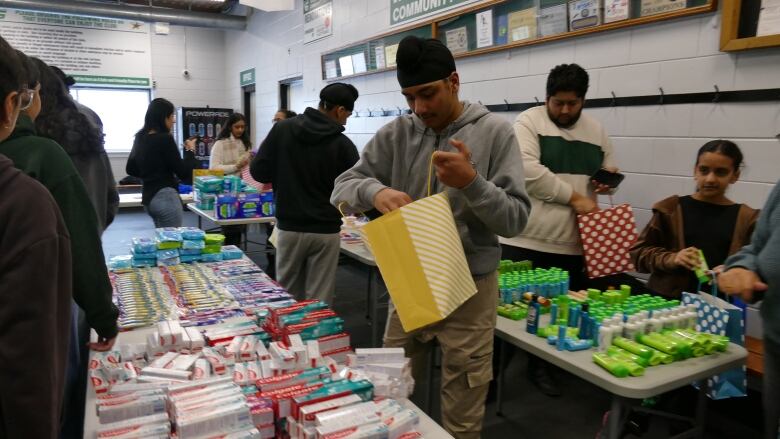 A group of people put together care packages. A boy is seen holding a bag in between two tables. One table is piled with packages of toothpaste, while the other is covered with deodorant and shampoo bottles.