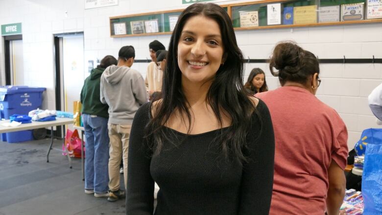 A woman smiles at the camera. A group of people behind her are lined up at a table, creating care packages.