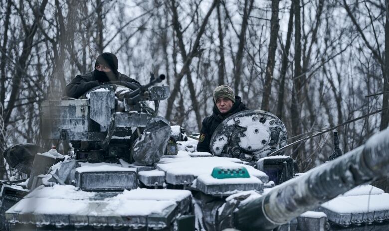 Two people are visible on top of a tank amidst trees. There is a light layer of snow on the tank.