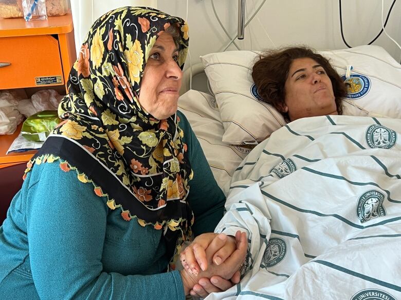 A woman with dark hair lies in a hospital bed, with a white blanket with blue-green stripes and circular logos pulled up to her neck. Next to her sits another woman, wearing a green shirt and a black, yellow and orange headscarf. The two hold hands as they look toward someone standing outside the photos frame.