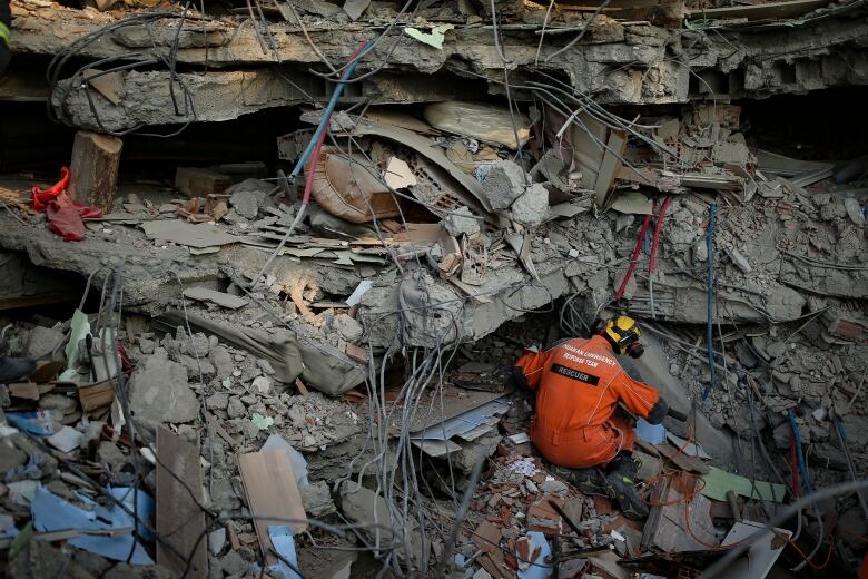 A rescue worker wearing an orange jumpsuit and yellow helmet searches a wall of concrete, mixed with wires, metal and people's belongings.