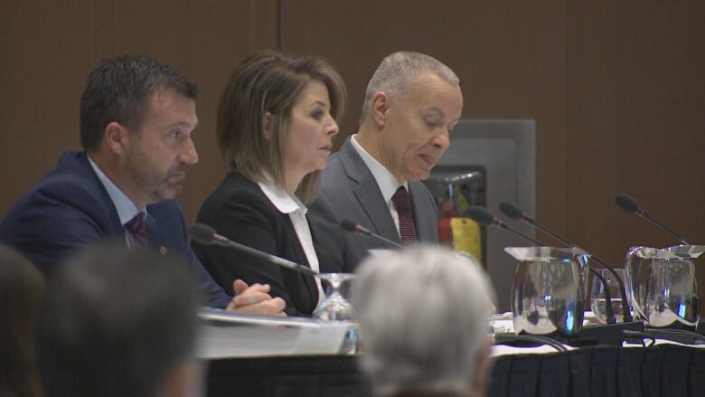 One woman and two men in suits sit behind microphones. 