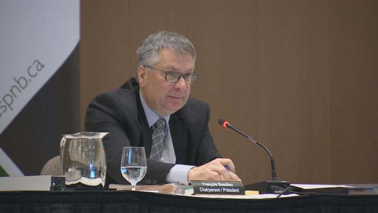 A man in a suit sits behind a table with a microphone. 