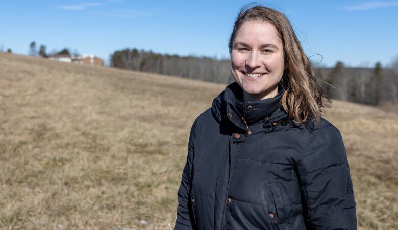 A white woman with shoulder-length brown hair smiles at the camera while standing outside in a field