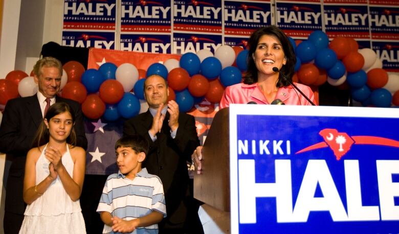 A woman is shown at a podium speaking into a microphone, with family members, campaign signs and balloons shown behind her.