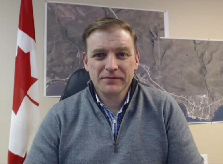 A person wearing a quarter zip sweater sits in front of a wall which has two maps. There is a Canadian flag to his left.