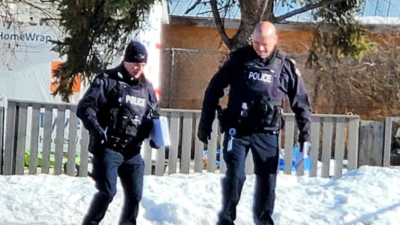 Two RCMP officers walk and talk on a sidewalk outside a white building bordered by a fence surrounding a snowy yard.