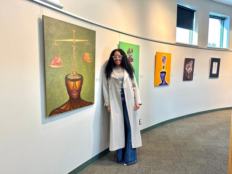 A painting shows part of a Black woman's face with a scale of justice balancing a heart and a brain on top of it. The artists stands in the gallery next to it.