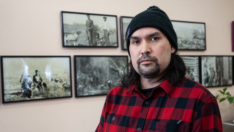 Dylan Loblaw, chief of the Ross River Dena Council, is pictured in a black toque and plaid shirt. He stands in front of a collection of photographs of elders.