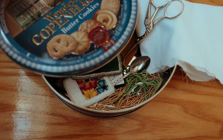 A cookie tin sits open. Inside is some beadwork, along with strings of beads and other supplies.