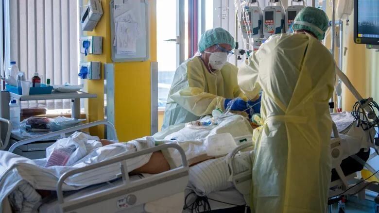 Two health-care workers care for a patient in a bed. The workers wear scrubs and personal protective equipment, including masks. 