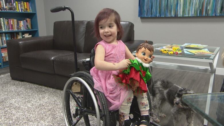 A little girl dressed in a pink frock sits with a doll in her wheelchair.