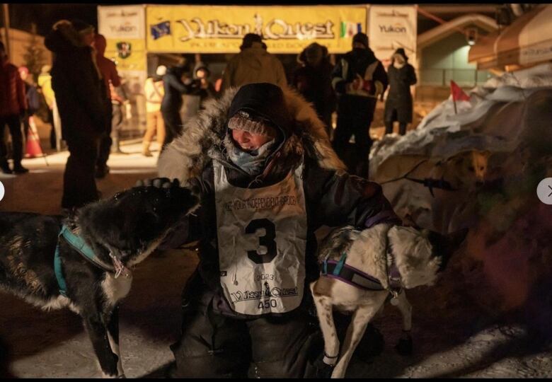 A woman in a parka and a bib with the number three kneels outside between two dogs.