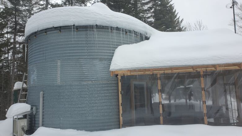 A grey farm silo sits covered in snow. Icicles hang from it's sides. It has a small enclosed porch on it's front.