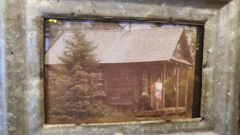 A framed photo of an old cabin with a woman standing on it's porch.