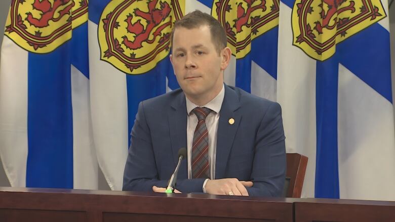 A man in a suit and tie is shown at a podium listening to questions from a reporter.