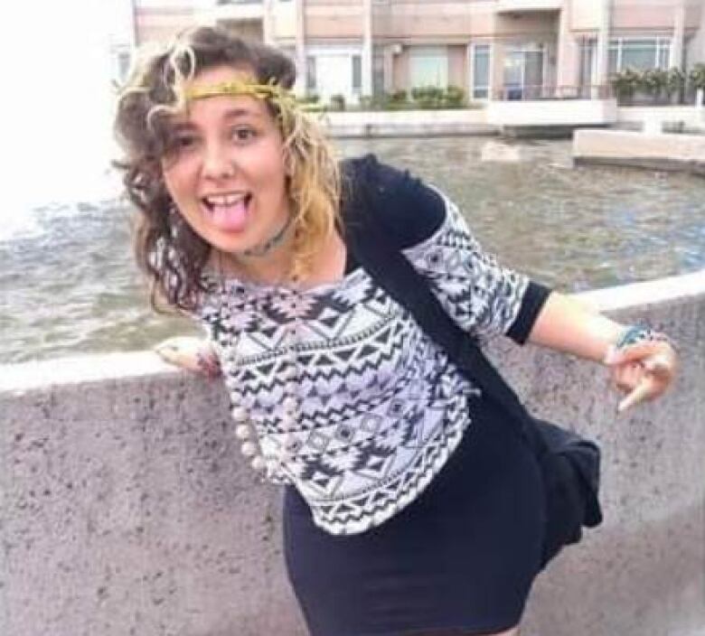 A girl in a black dress and a white sweater is shown in front of a fountain, with a look of glee on her face.