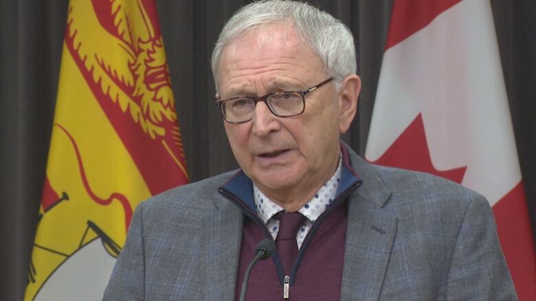 An older man with a furrowed brow standing in front of a New Brunswick flag and a Canada flag.