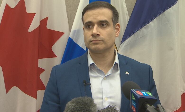 A man with short dark hear and wearing a blazer stands in front of Canada and Nova Scotia flags.