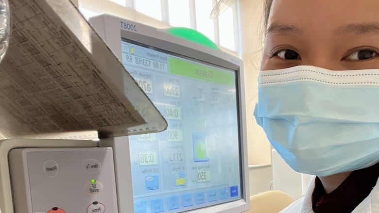 A nurse wearing a mask stands beside a medical monitor.