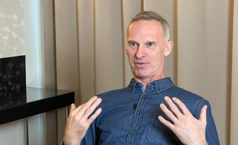 A man in a blue shirt with blue eyes sits looks incredulous or exasperated as his hands gesture in front of his chest. 