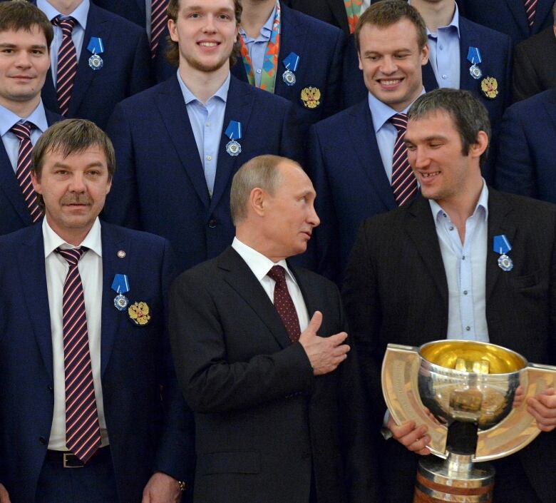 A group of men in dark suits stand for a picture, some with ties some without. Front and centre, the only man without a medal, Russian president Vladimir Putin, looks up at and gestures to a tall man with the medal, holding a large gold cup. The medal is blue and pinned on the left lapel. 