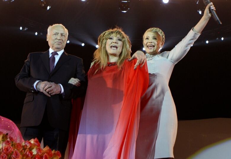 Three people stand on a stage, with flowers at their feet. A woman in a red and pink dress with long hair is in the middle, with a woman in a white dress to her right in the picture, and a man in a black suit to her left. 