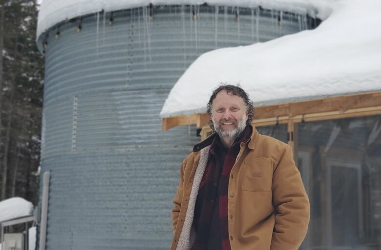 A man with a greying beard stands smiling wearing a tan jacket over a plain shirt in front of a silver grain silo covered in snow