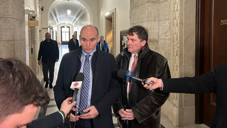 Two men stand side-by-side speaking into microphones held in front of them. One man, at left, is bald and wearing a blue shirt and striped tie. The other man, at right, has brown hair and is wearing a brown coat.