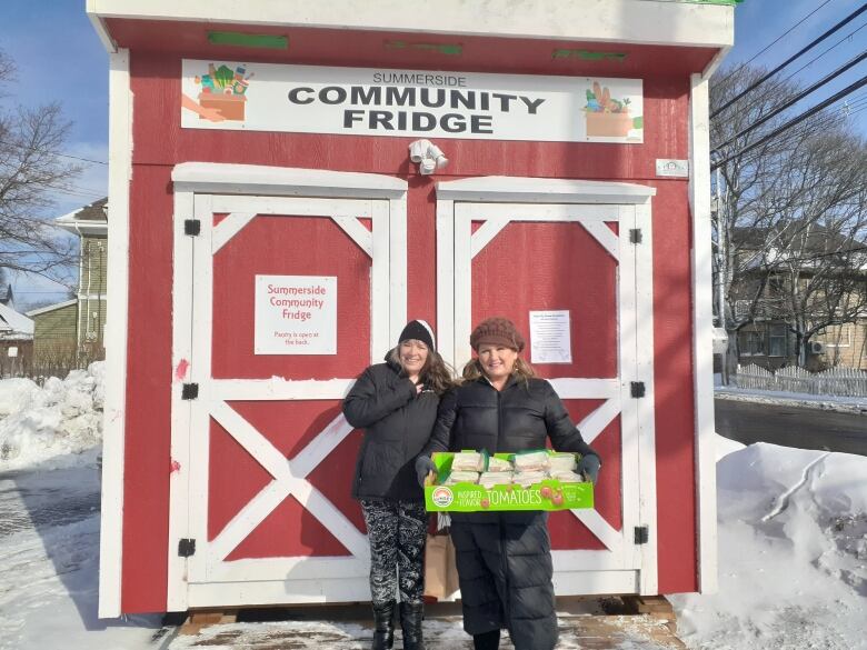 Two women stand in front of a red shed with two doors. One holds a carboard pallet of sandwiches. 