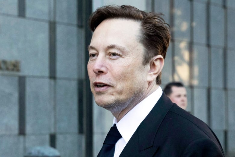 Man with brown hair and wearing suit and tie walks outside an office building and looks in the distance.