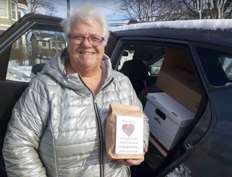 A woman in a silver parka and white hair holds a brown paper bag lunch. A label on front says Made with Love.