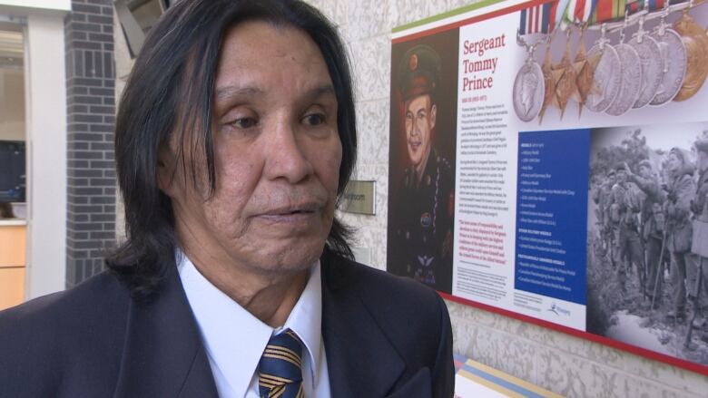 A man with shoulder-length black hair wearing a suit stands in front of a display of Sgt. Tommy Prince.