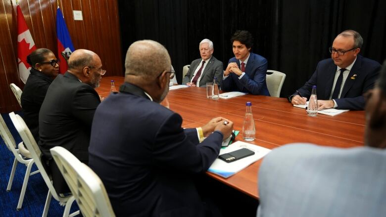 Six men wearing suits sit around a boardroom table and talk, a Canadian flag and the flag in Haiti in background