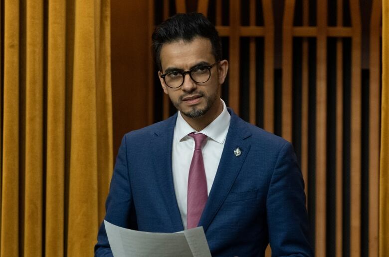 An Arab man speaking at the House of Commons in a navy blue suit and pink tie with round black glasses holding a piece of paper 