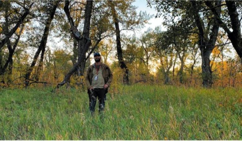 A man wearing a green sweater stands in a field, there are autumn leaves on the trees behind him